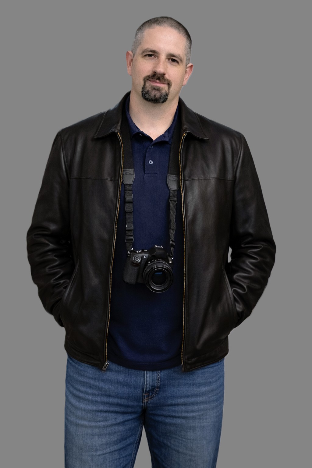 Man with short hair and a beard wearing a black leather jacket, blue jeans, and a camera around his neck, standing against a plain gray background.
