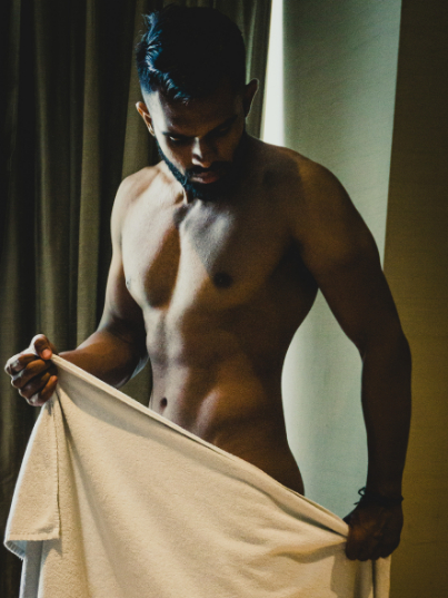 Shirtless man standing indoors, holding a white towel around his waist, looking down.