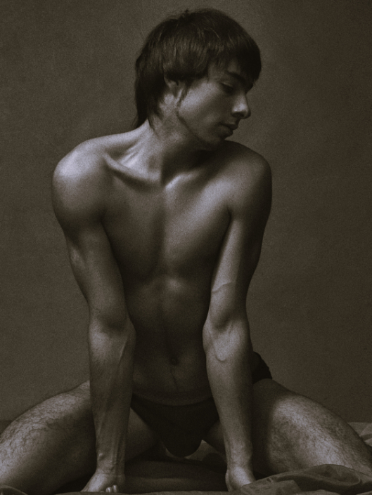 A shirtless man sits on his knees, arms extended downward, looking over his left shoulder against a plain background.